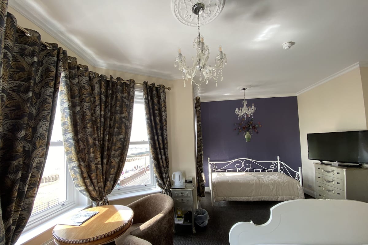 A hotel room with a bed, a TV, a chandelier, a window with curtains, a small table, and a chair.