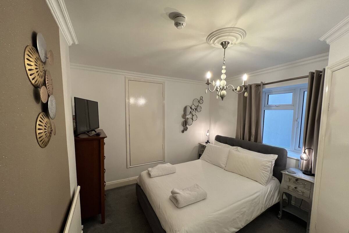 A neatly arranged bedroom with a bed, a television on a wooden cabinet, a ceiling fan, a chandelier, and a window with curtains.