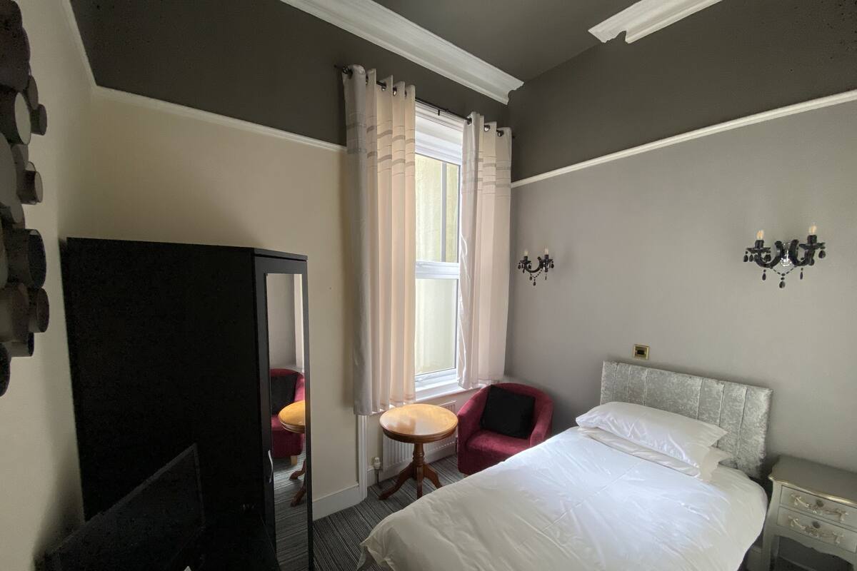 A hotel room with a bed, a chair, a small table, a mirror, and a nightstand.