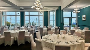 A dining area with round tables, white tablecloths, and chairs, set against a backdrop of large windows with a view of the sea. - The Cumberland Hotel (Eastbourne)