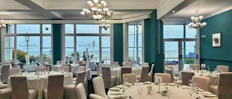 A dining area with round tables, white tablecloths, and chairs, set against a backdrop of large windows with a view of the sea.