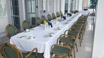 A long dining table with white tablecloths, surrounded by wicker chairs with green cushions, set for a meal.