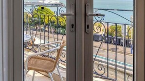 A balcony with a wicker chair, a metal railing, and a view of the sea and a building in the distance. - The Cumberland Hotel (Eastbourne)