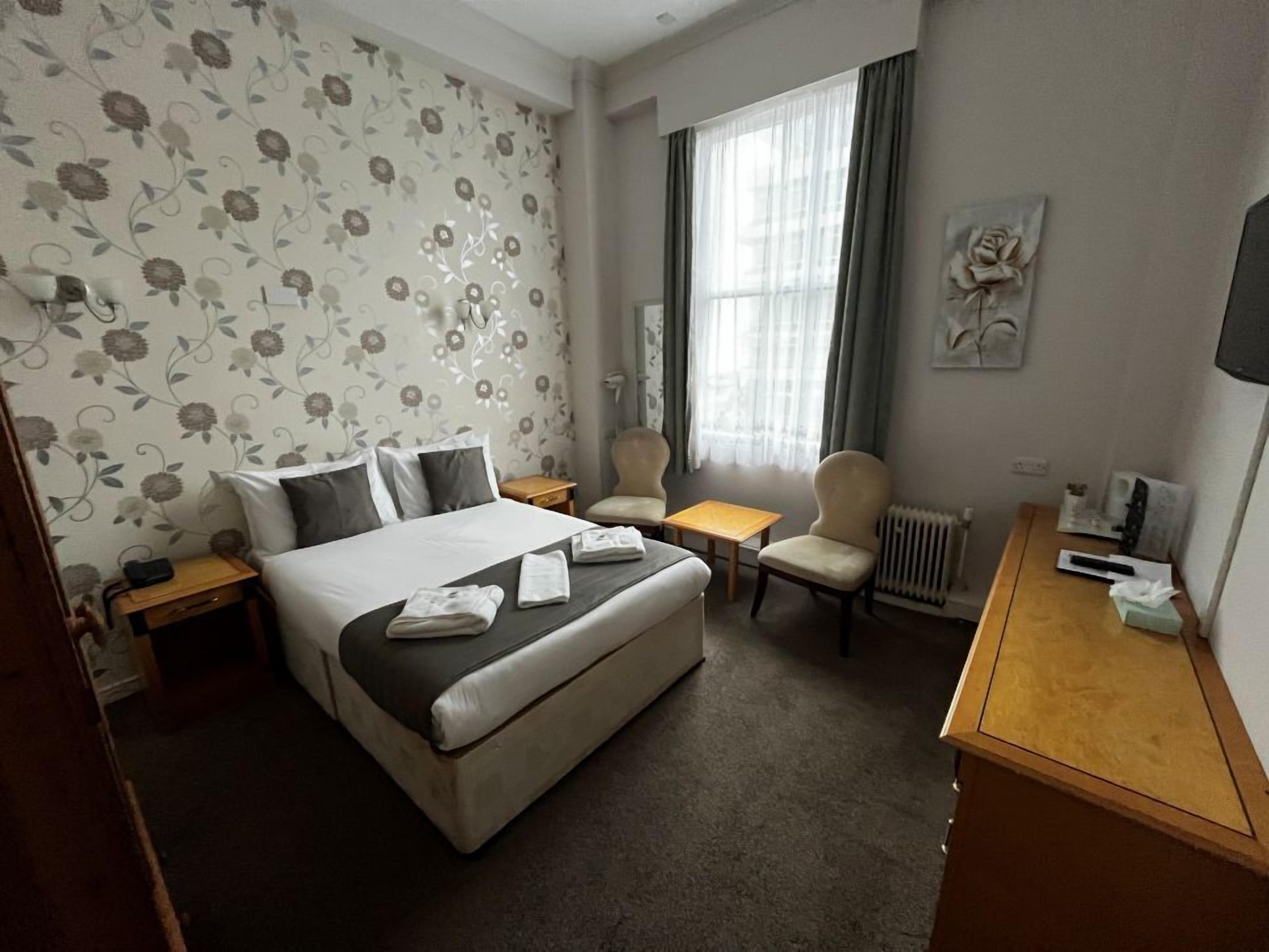 A hotel room with a bed, two chairs, a small table, a radiator, and a TV.