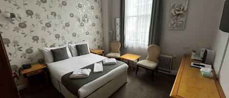A hotel room with a bed, two chairs, a small table, a radiator, and a TV.