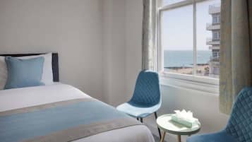 A hotel room with a bed, two chairs, a small table, and a window with a view of the beach and buildings.