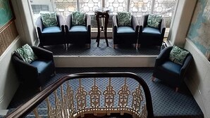 Een zithoek met twee banken en een kleine tafel, een decoratieve balustrade en uitzicht door een raam met een datumbord.