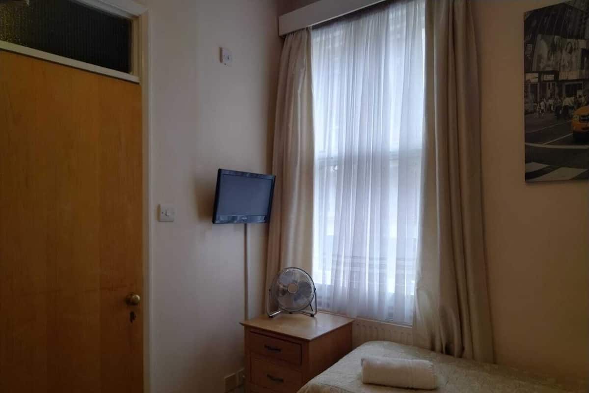 Une chambre avec une armoire en bois, une table de chevet avec un ventilateur, un téléviseur fixé au mur et une fenêtre ornée de rideaux légers.