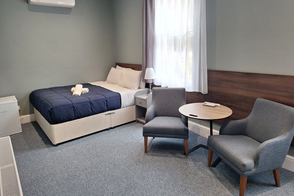 Une chambre d’hôtel avec un lit, deux fauteuils, une petite table, une lampe et une fenêtre avec des rideaux.