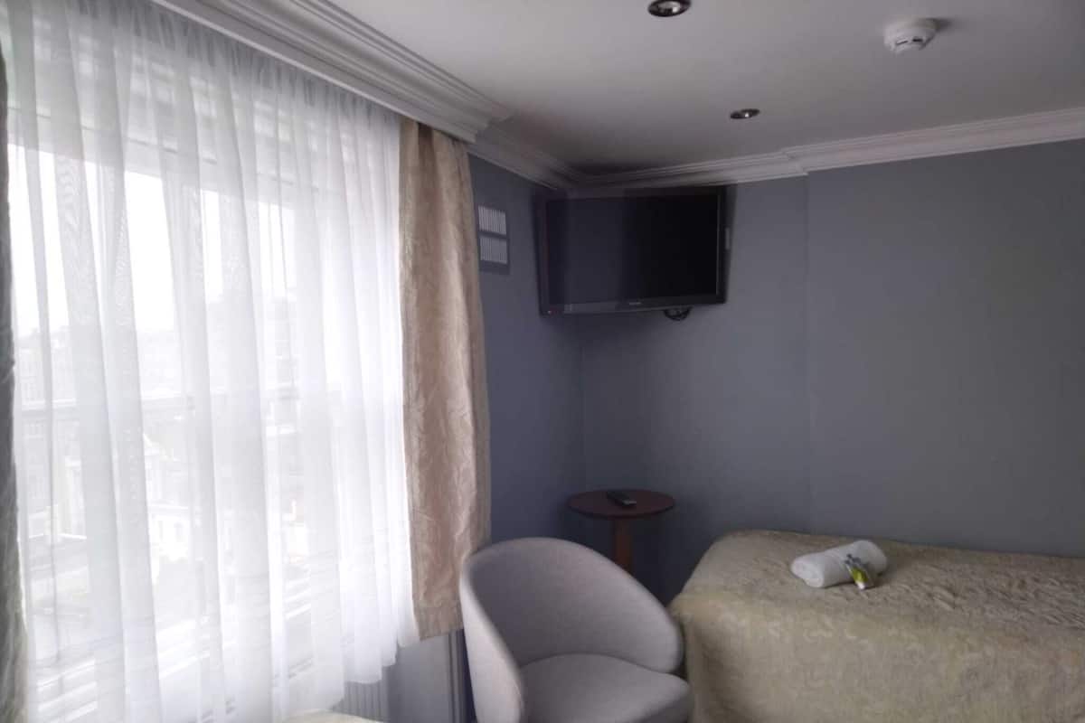 Une chambre d’hôtel avec un lit, une chaise, une petite table, un téléviseur fixé au mur et une fenêtre avec des rideaux légers.