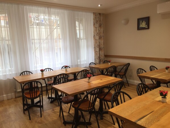 A dining area with wooden tables and chairs, large windows with sheer curtains, and a framed picture on the wall. - St Simeon (London)