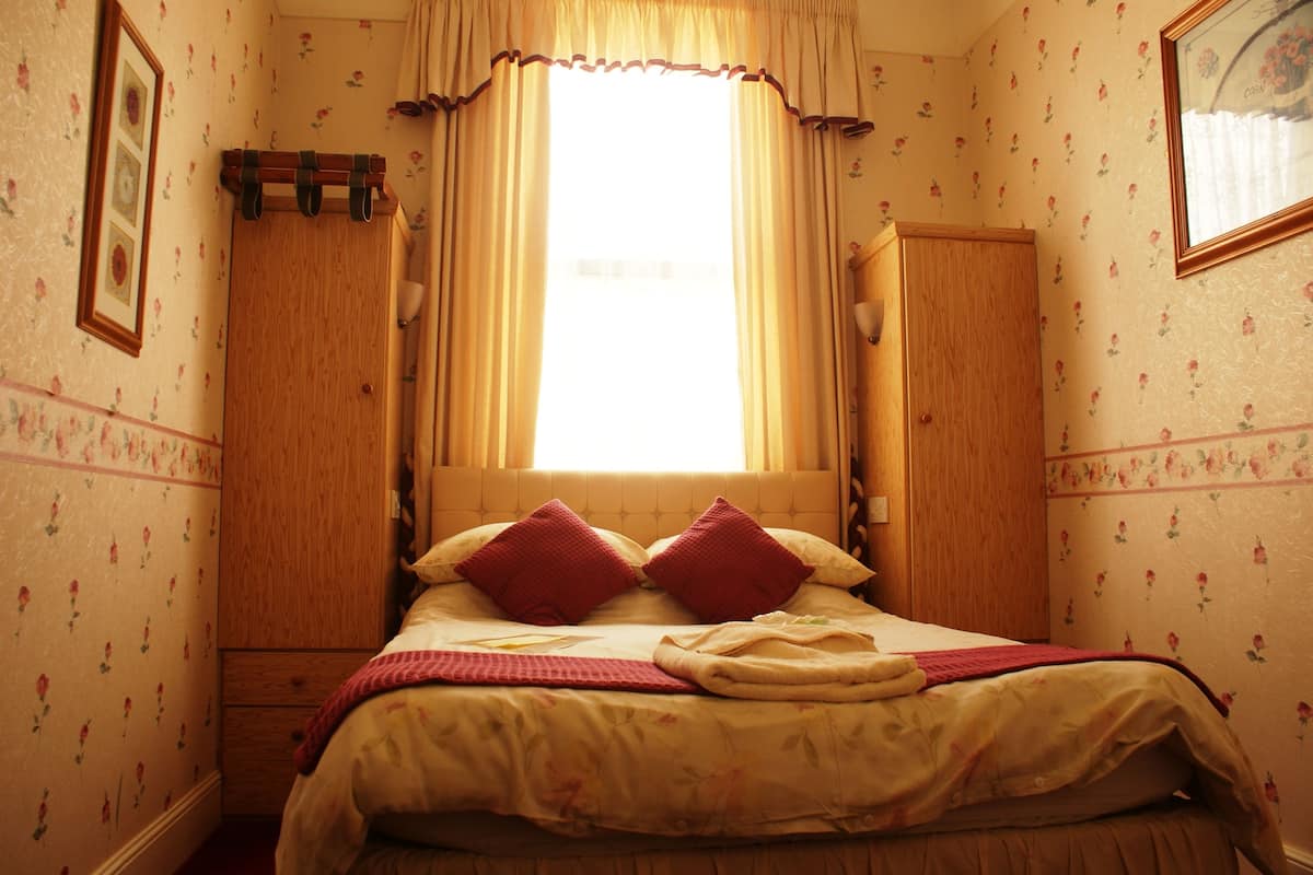 A bedroom with a bed, a window with curtains, a wooden wardrobe, and floral wallpaper.