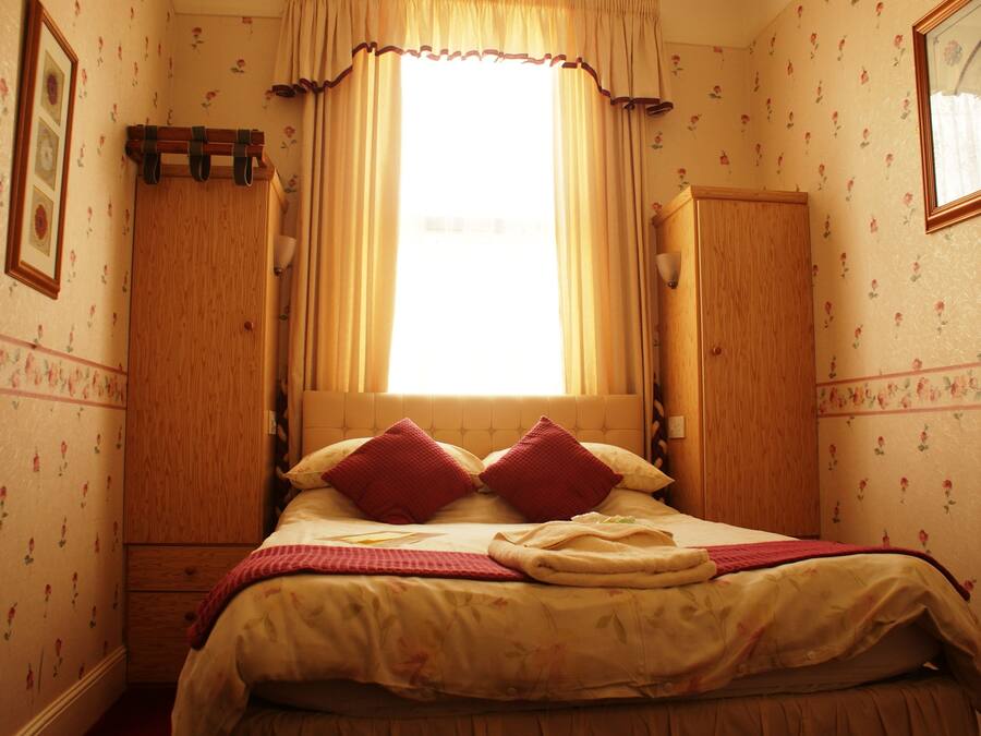 A bedroom with a bed, a window with curtains, a wooden wardrobe, and floral wallpaper.