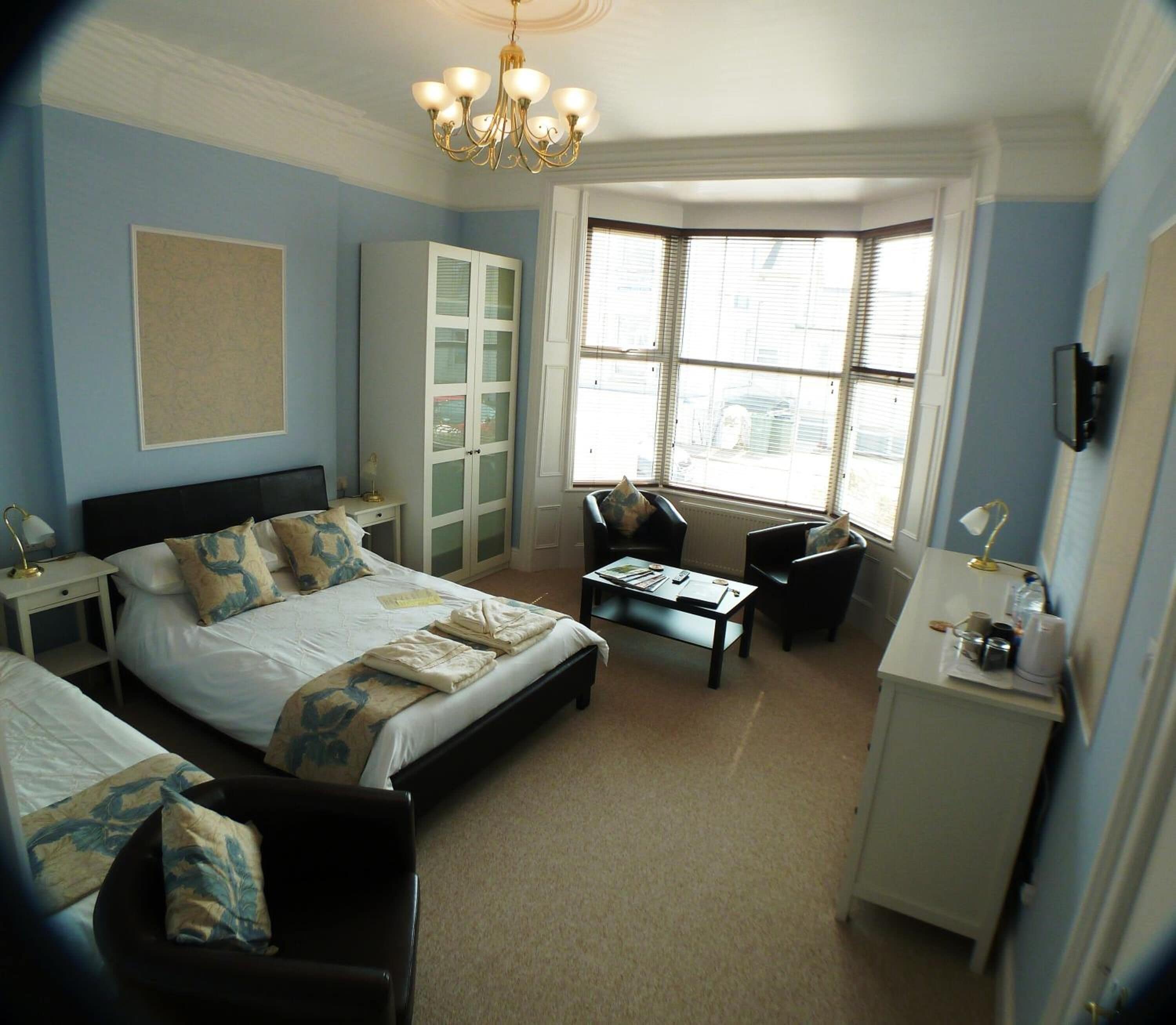 A bedroom with a large bed, a desk, and a chair. There is a wardrobe and a window with blinds.