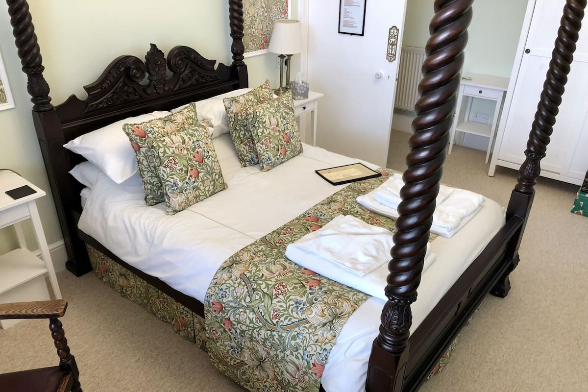 A four-poster bed with floral bedspread and pillows, a bedside table with a lamp, and a framed certificate on the bed.