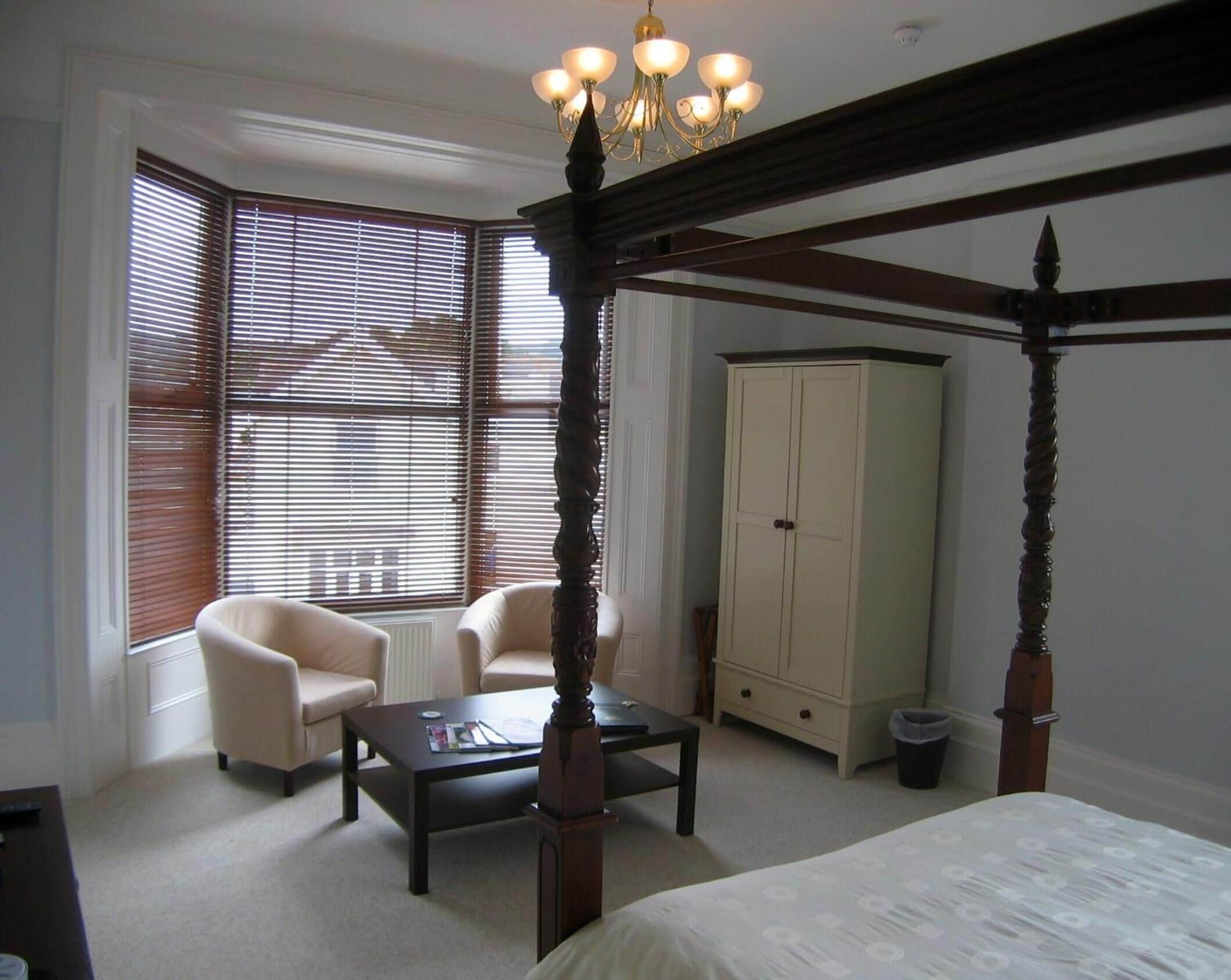 A bedroom with a four-poster bed, a wooden desk, a chair, a cabinet, and a window with blinds.