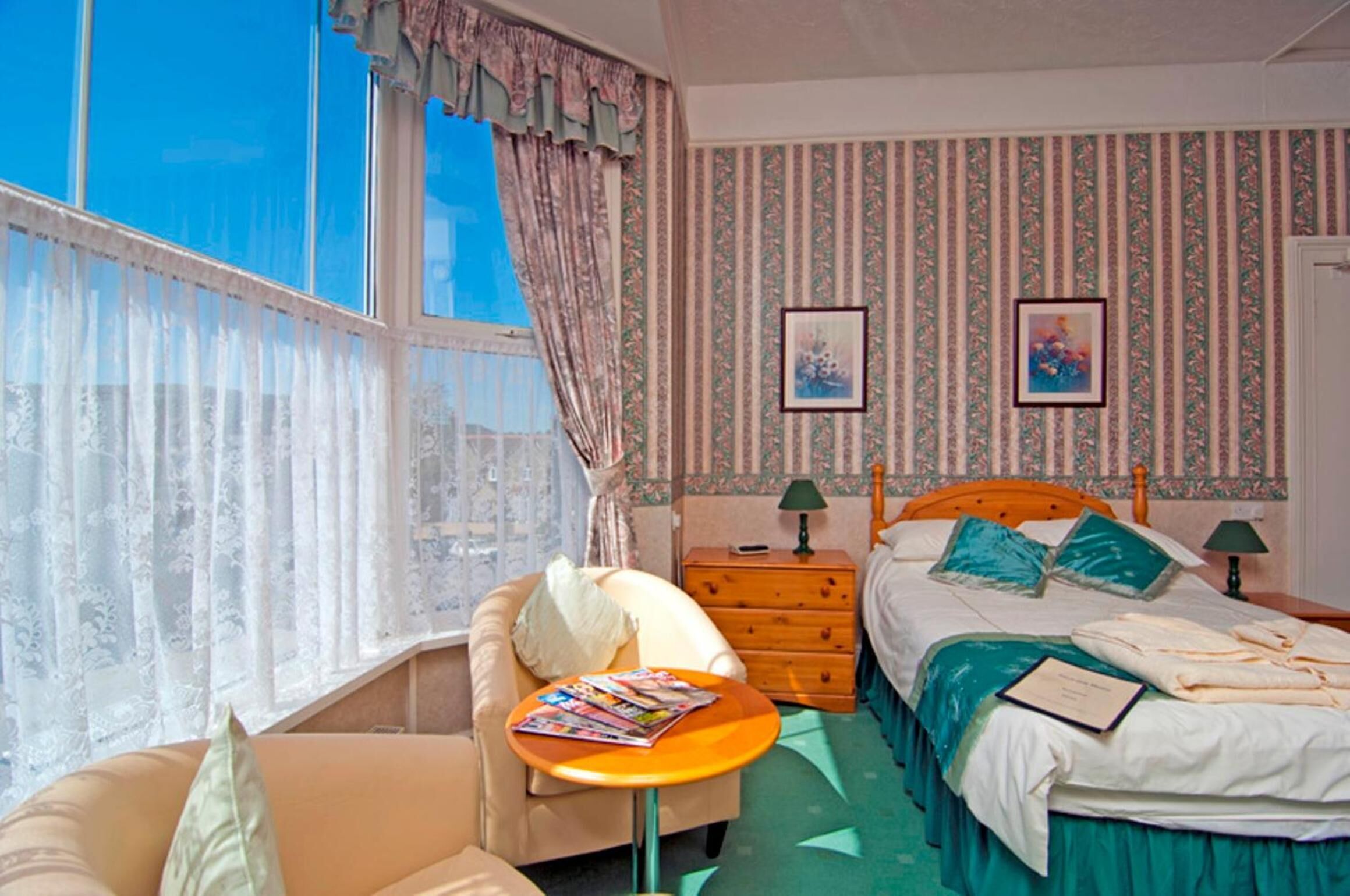 Une chambre d’hôtel avec un lit, une chaise, une petite table, une table de chevet et une fenêtre avec des rideaux.