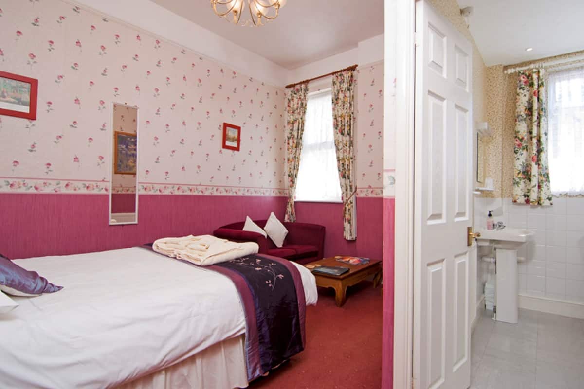 A bedroom with a bed, a red armchair, a small table, and a bathroom with a sink and mirror.