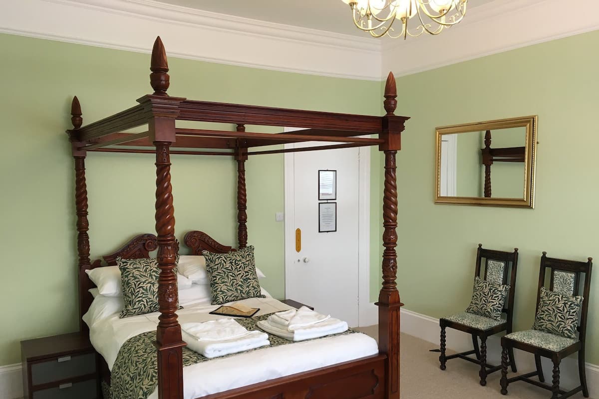 A four-poster bed with a canopy, two upholstered chairs, a mirror, and a door with framed certificates.