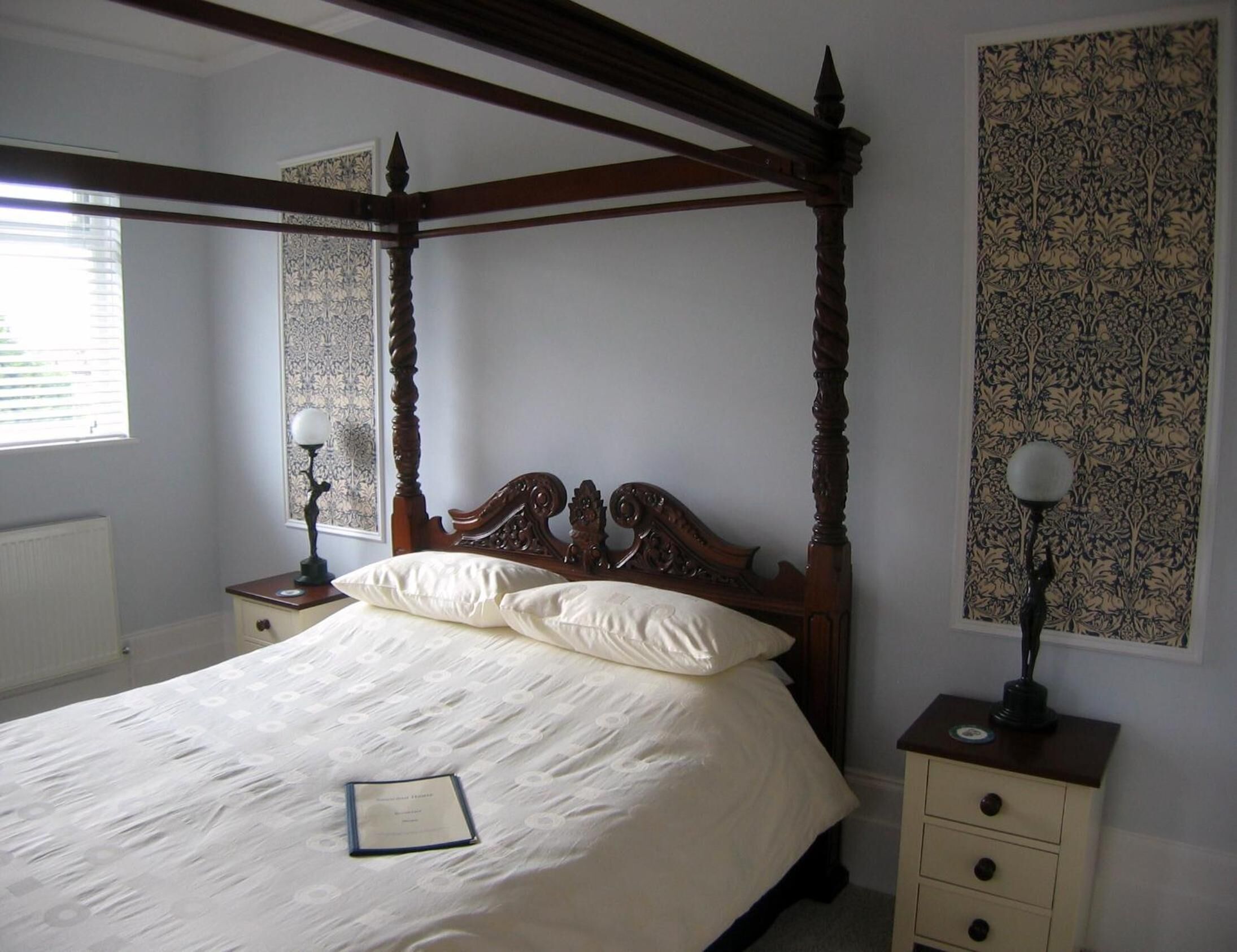Una cama con dosel, un cabecero de madera, dos mesitas de noche con lámparas y un papel tapiz estampado en las paredes.