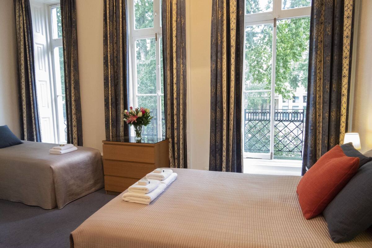 Ein Hotelzimmer mit einem großen Bett, einem kleineren Bett, einem Schminktisch und einem Fenster mit Vorhängen.