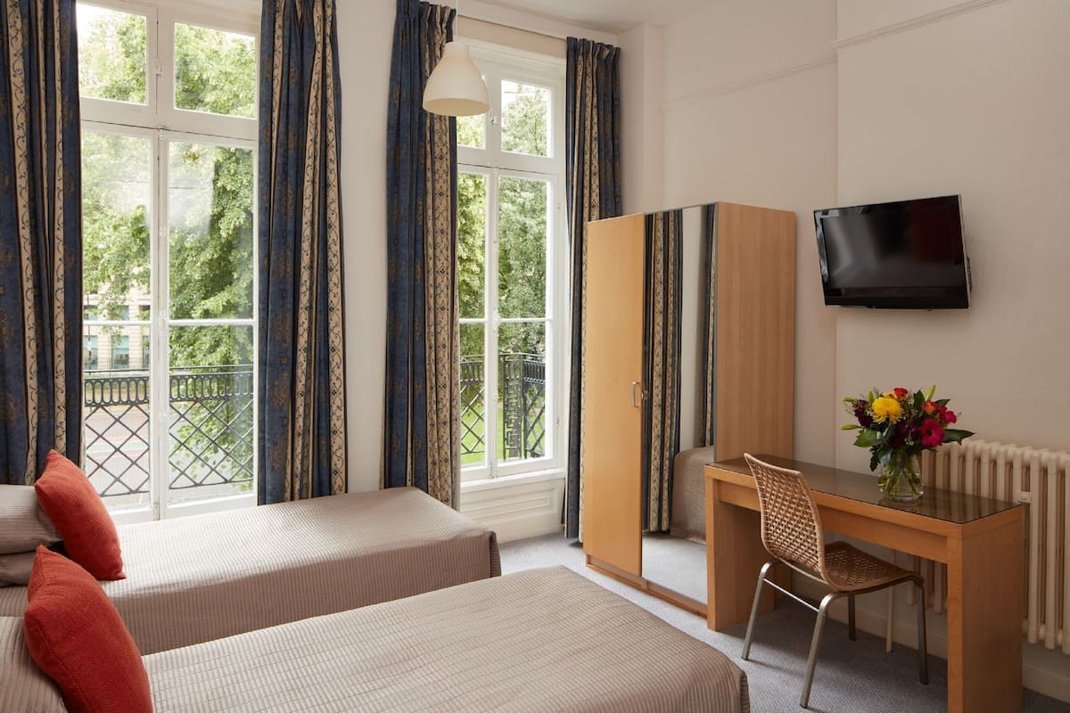 Ein Hotelzimmer mit zwei Betten, einem Schreibtisch, einem Stuhl, einem Fernseher und einem Fenster mit Vorhängen.