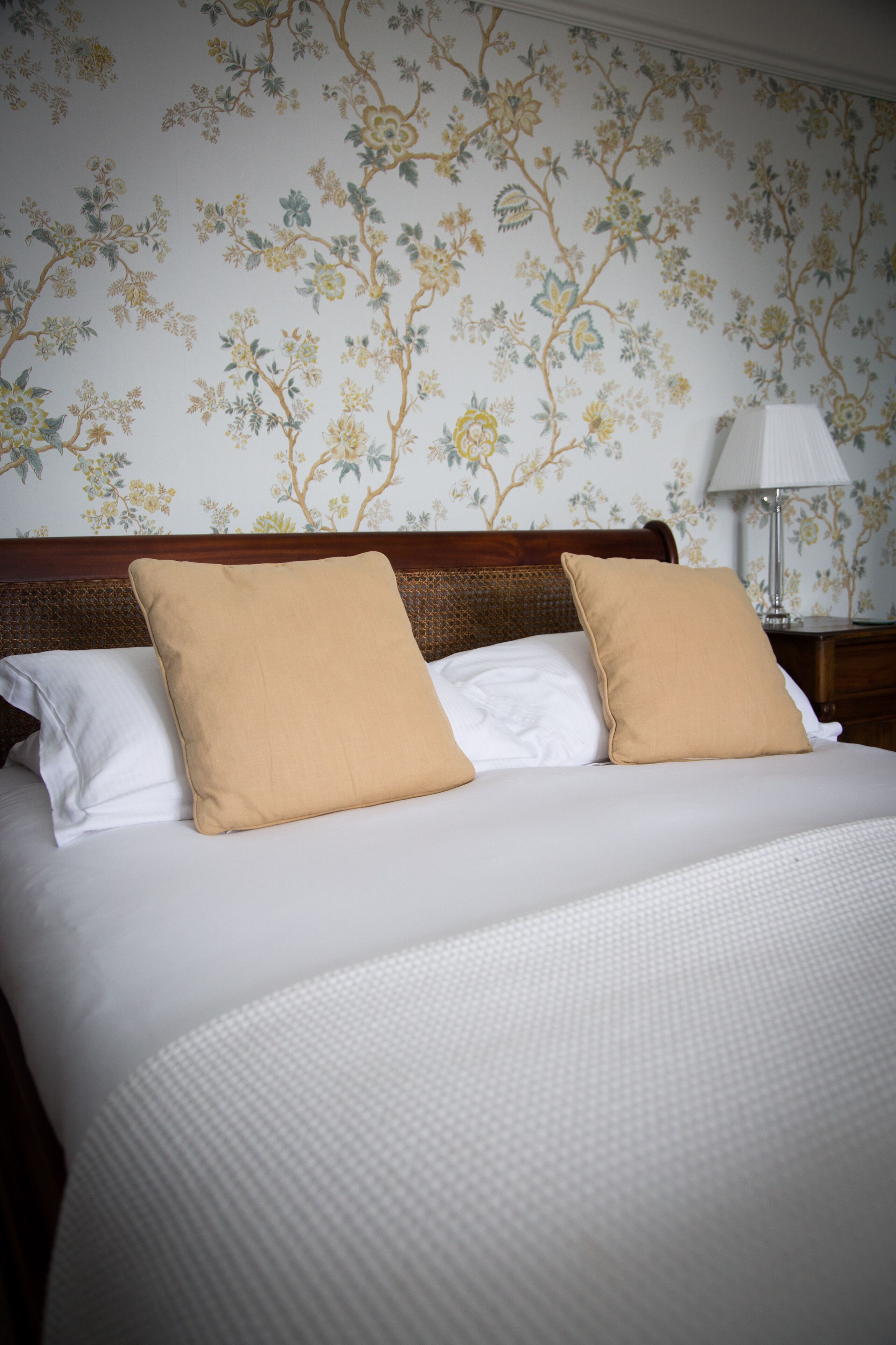 Een net opgemaakt bed met witte en beige kussens, een houten hoofdbord en een behang met een bloemenpatroon.