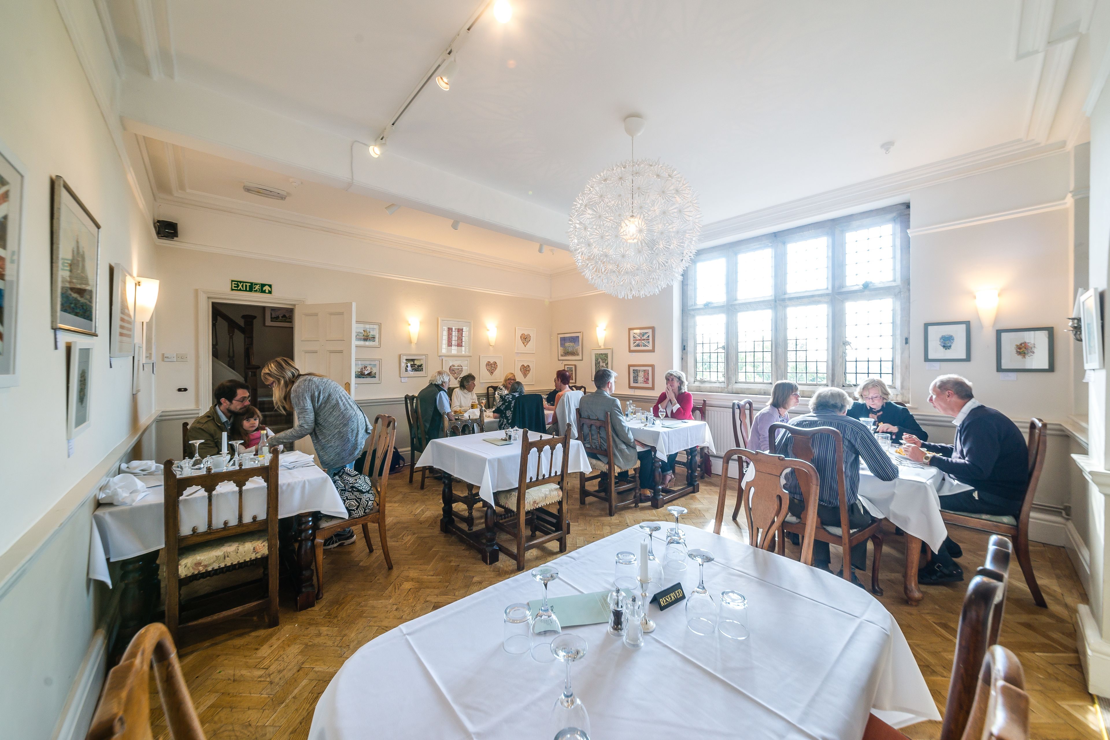Un espace repas comprenant plusieurs tables, des chaises et un lustre. Les murs sont ornés de tableaux encadrés et un panneau indiquant la sortie est visible.