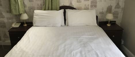 Una cama bien hecha con sábanas blancas, dos mesitas de noche con lámparas, y una pared con papel tapiz con motivos parisinos.
