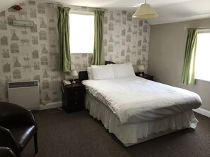 Siististi järjestetty hotellihuone, jossa on suuri sänky, kaksi yöpöytää valaisimineen, tuoli ja patteri.