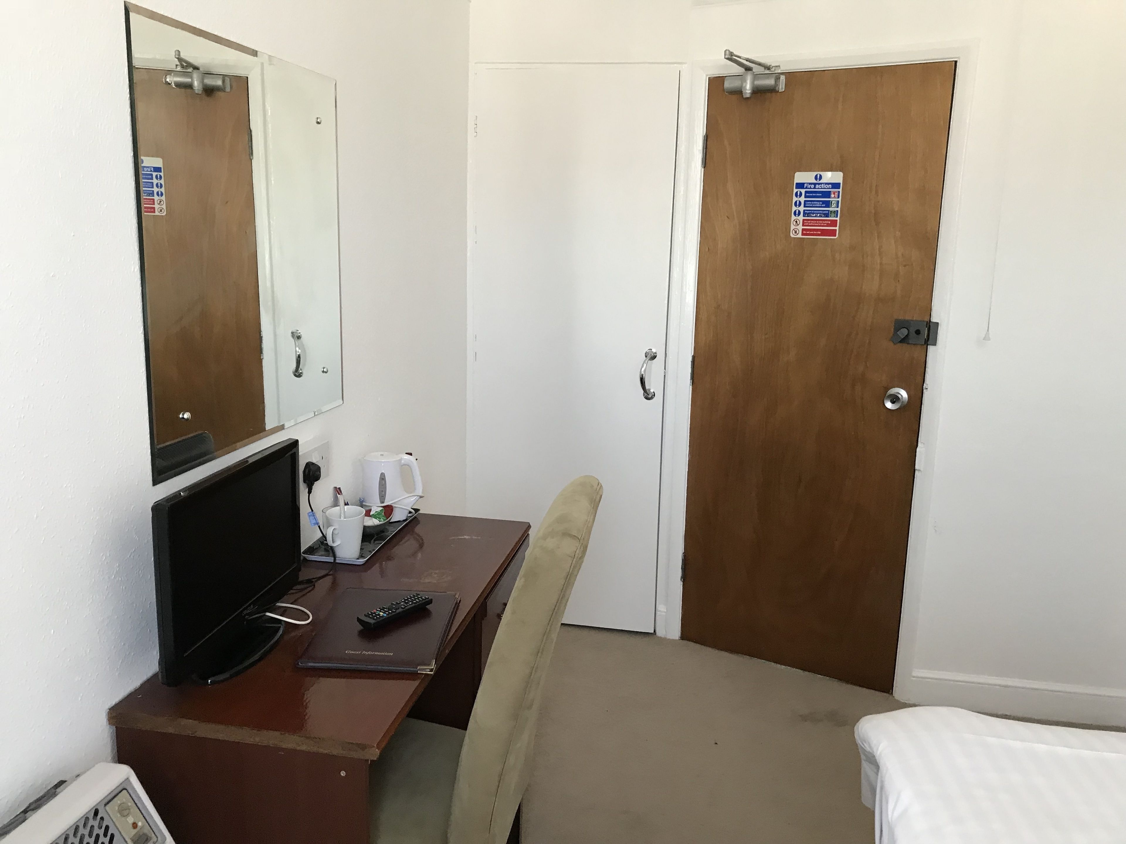 A hotel room with a wooden desk, a chair, a bed, a TV, and a mirror. There are signs on the doors indicating fire action and no smoking.