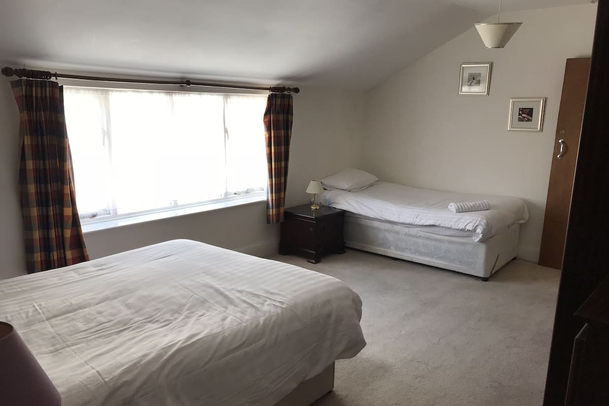 A bedroom with a large bed, a smaller bed, a nightstand, a lamp, and a window with curtains.