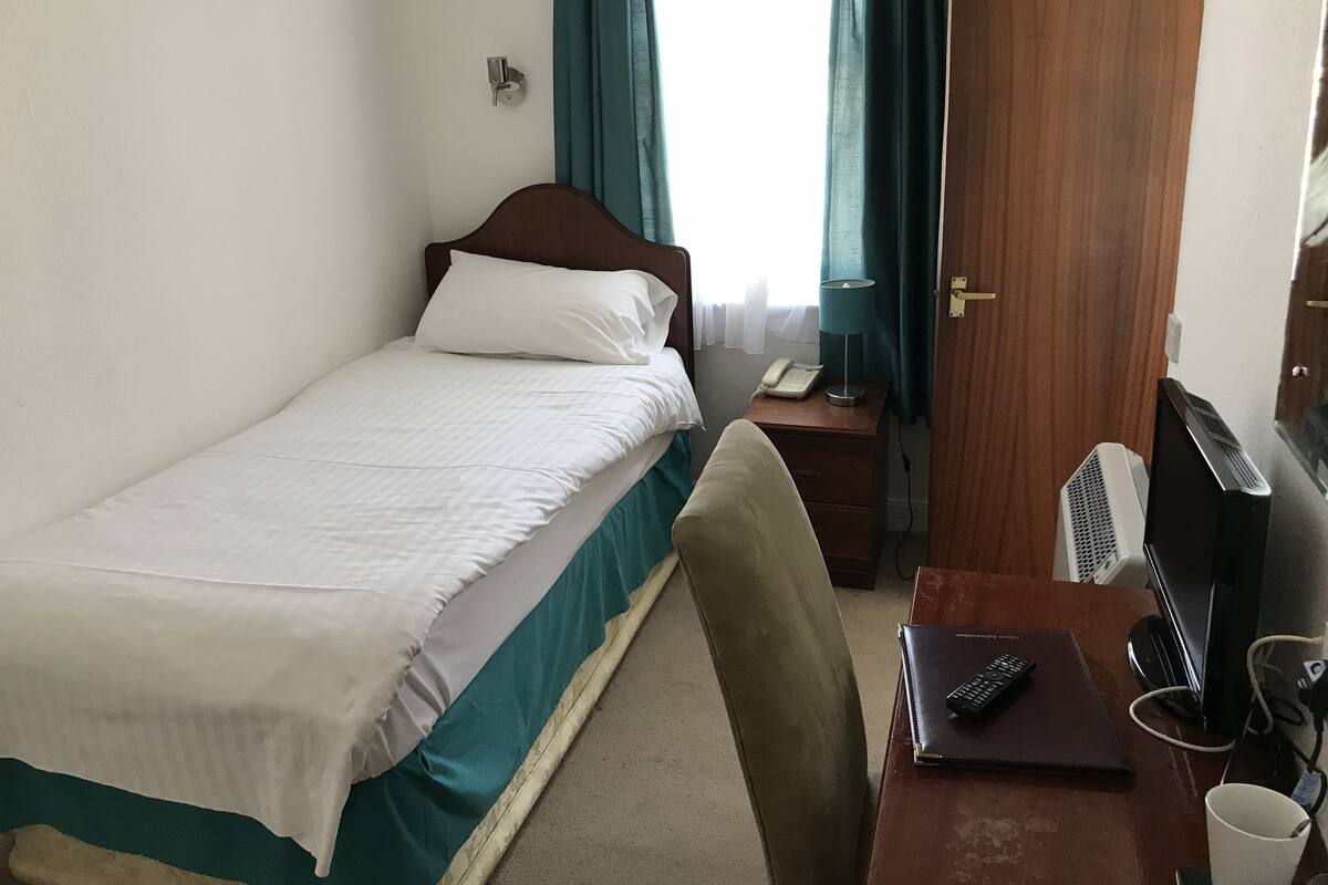 A single bed with white bedding, a bedside table with a lamp, a desk with a computer and a remote control, and a door with a wooden frame.