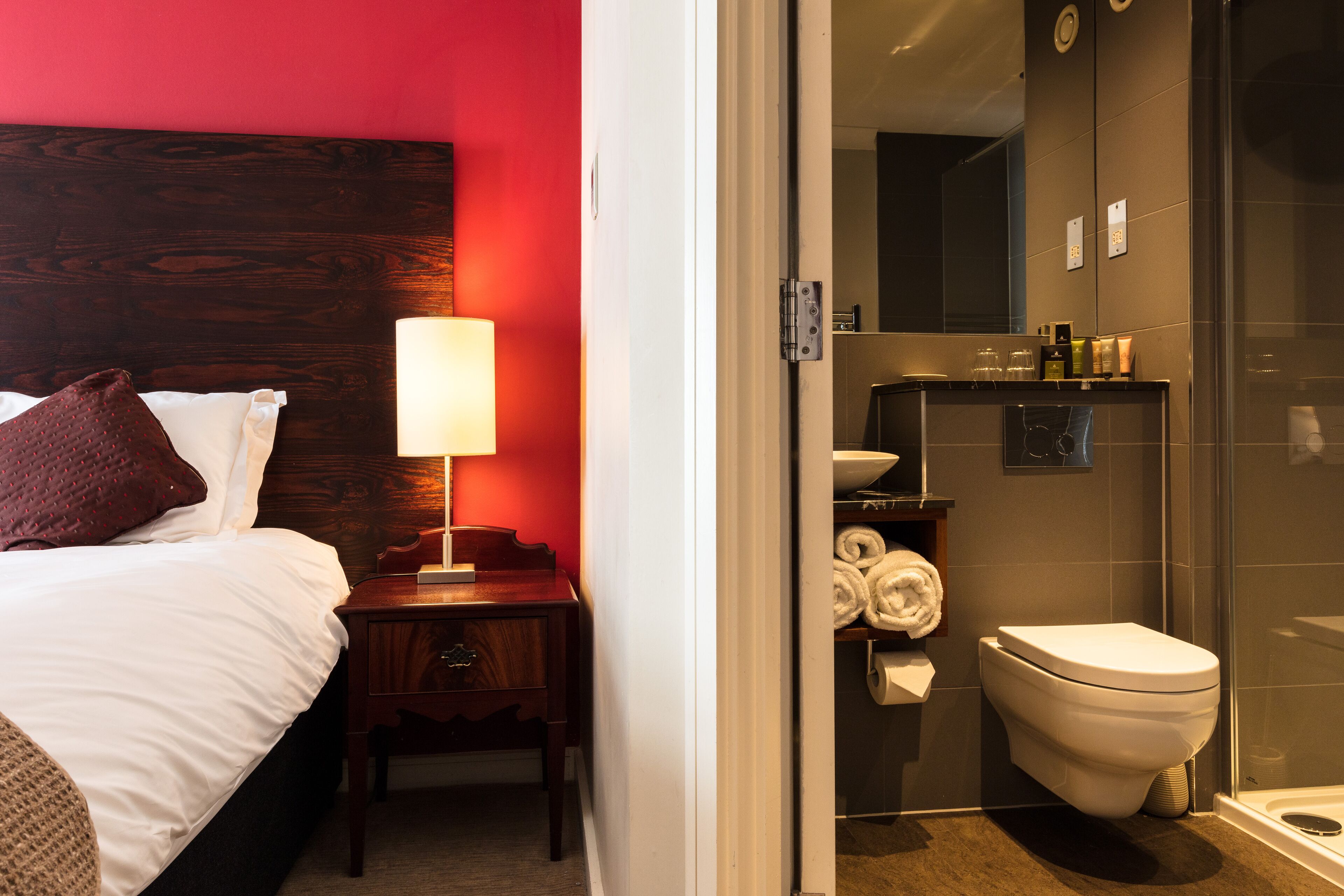 Une chambre d’hôtel comprenant un lit, une table de chevet, une lampe et donnant sur la salle de bain, qui est équipée de toilettes, d’un porte-serviettes et d’une douche.