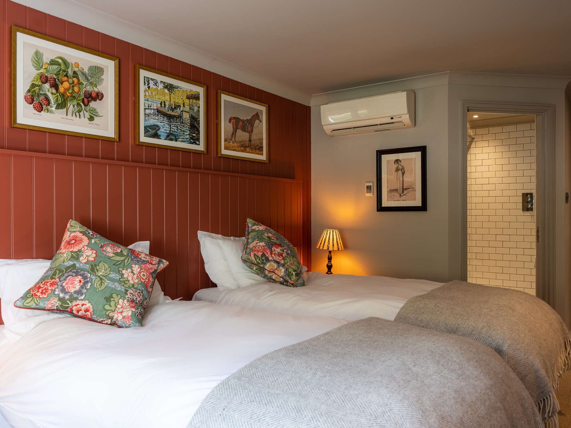 Habitación de hotel con dos camas, almohadas estampadas con motivos florales, cuadros enmarcados con botánica y paisajes, un equipo de aire acondicionado y una pared de ladrillo.