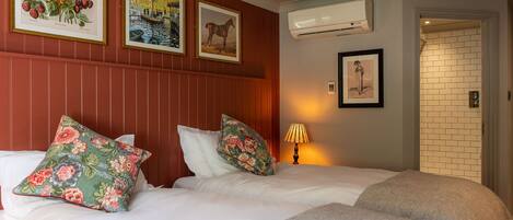 Habitación de hotel con dos camas, almohadas estampadas con motivos florales, cuadros enmarcados con botánica y paisajes, un equipo de aire acondicionado y una pared de ladrillo.