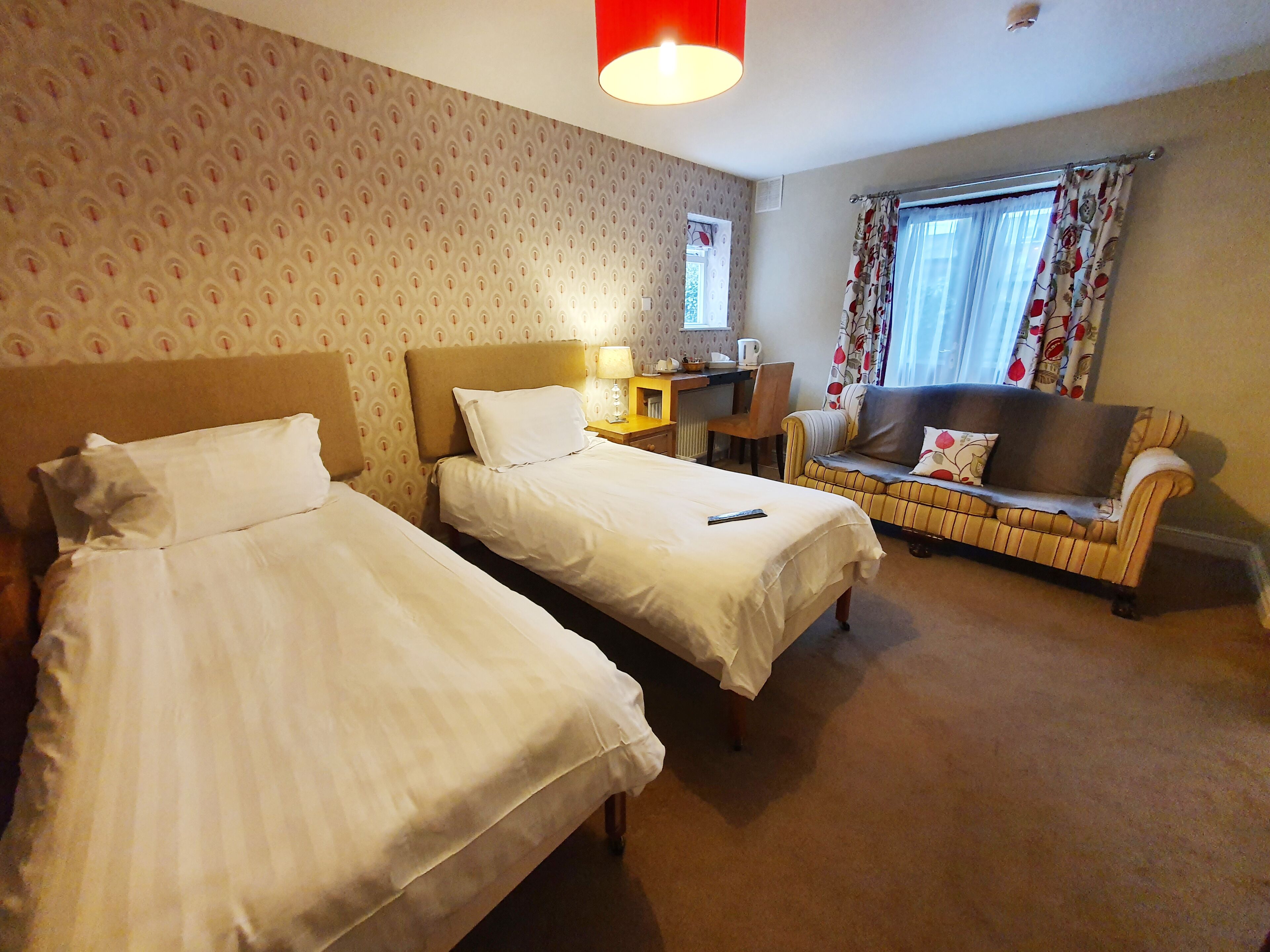 A hotel room with two single beds, a sofa, a desk with a lamp, and a window with curtains.