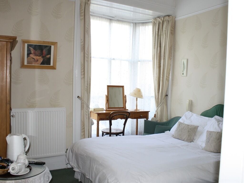 A hotel room with a large bed, a desk with a chair, a radiator, and a window with curtains.