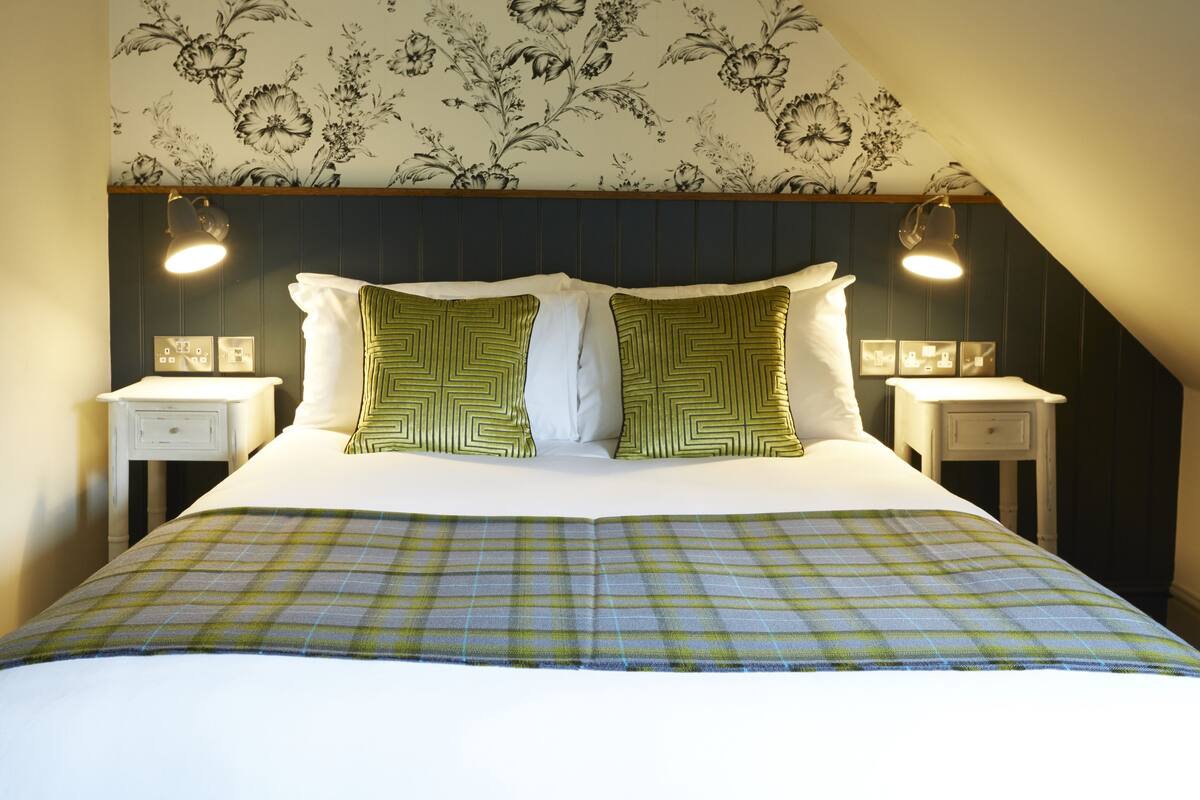 A neatly made bed with a plaid bedspread, two green patterned pillows, and two bedside lamps on matching nightstands.