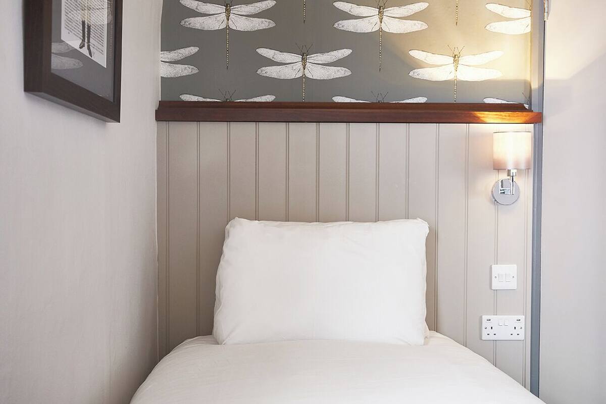 A single bed with a white pillow and a grey blanket, a wall-mounted light fixture, and a framed picture on the wall.