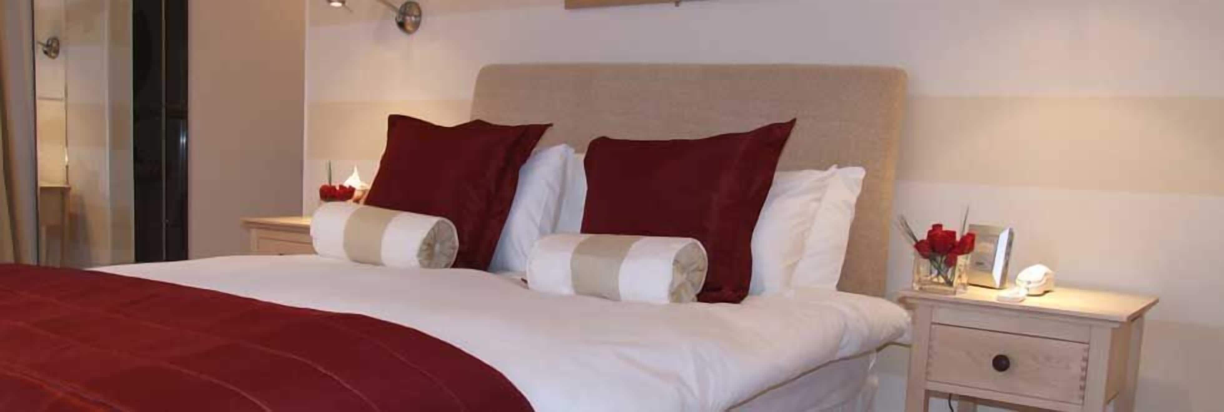 Una cama bien hecha con una colcha roja, almohadas blancas y un cabecero color beige. Hay una mesita de noche con una lámpara y un jarrón con rosas rojas.