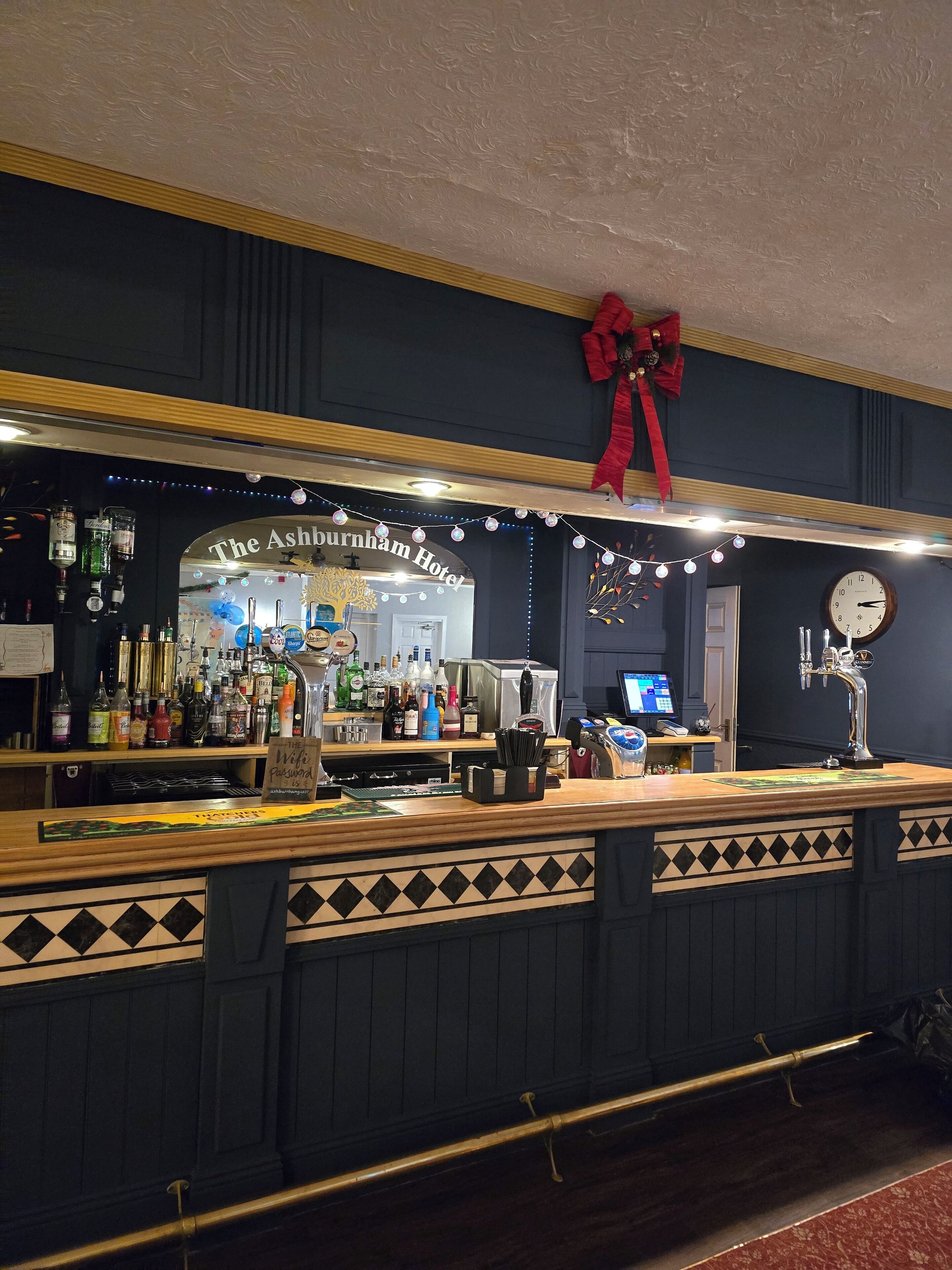 Eine Bar mit einer Theke, Regalen voller Spirituosenflaschen, einer Uhr und einem Schild für The Ashburnham Hotel.
