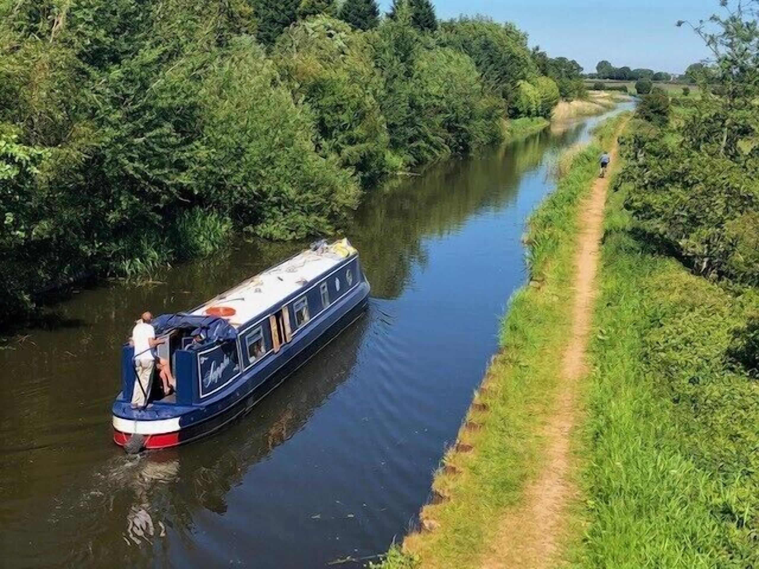 Um canal estreito com um barco fluvial atracado à esquerda, rodeado por vegetação densa e um caminho de terra à direita.