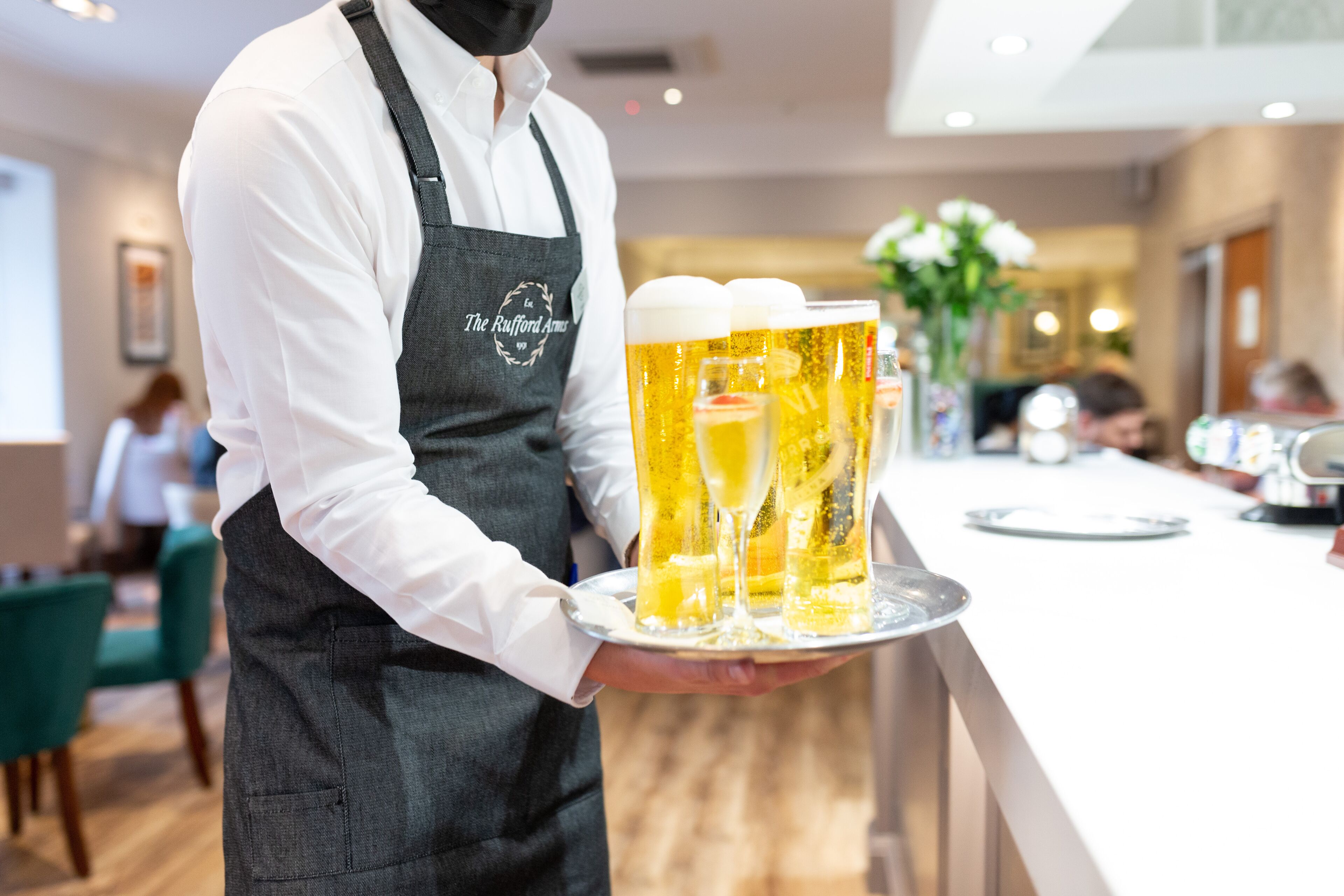 Un cambrer amb camisa blanca i davantal negre portant una safata amb tres gots de cervesa.