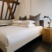 Un dormitorio moderno con una cama grande, varios almohadones, una mesita de noche, una lámpara y un cabecero de madera.