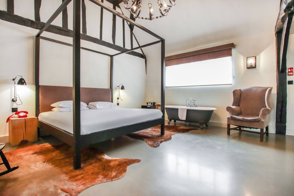 A modern bedroom with a four-poster bed, a vintage-style bathtub, a leather armchair, and a wooden nightstand with a lamp.