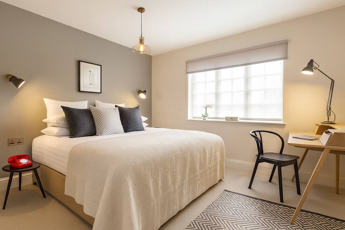 A modern hotel room with a large bed, a desk, and a chair. There is a window with blinds, a wall-mounted telephone, and a decorative picture frame.