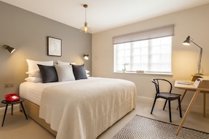 Una habitación de hotel moderna con una cama grande, un escritorio y una silla. Tiene un ventanal con persianas, un teléfono instalado en la pared y un marco decorativo.