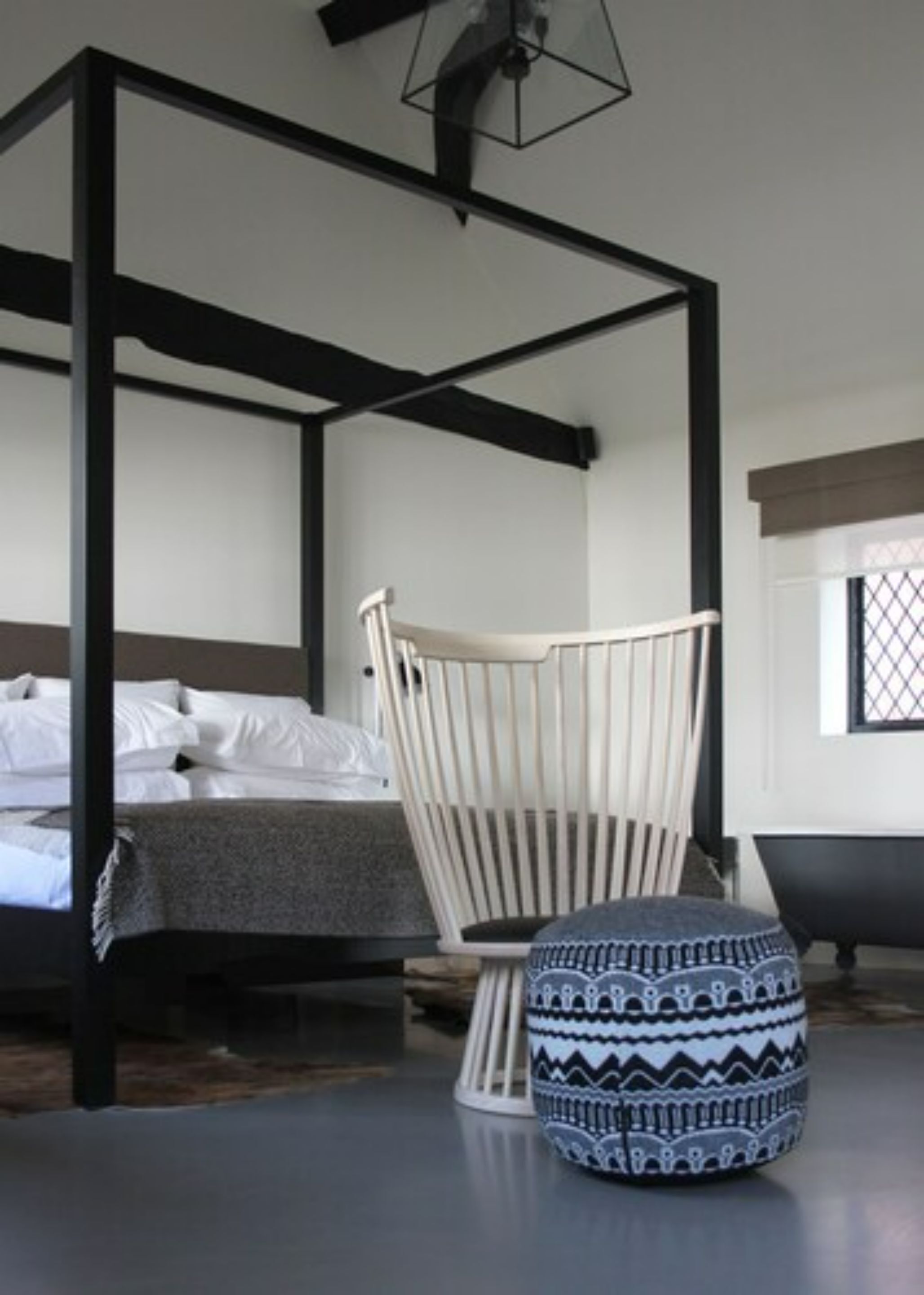 Ein Bett mit Baldachin mit weißen Leinen, ein weiß gepolsterter Sessel und ein gemusterter Hocker in einem modernen Schlafzimmer mit dunklem Boden und einem Fenster mit Gittermuster.