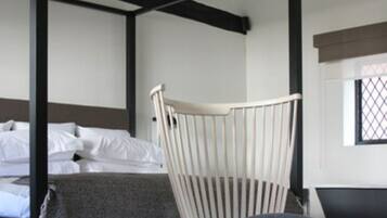 Ein Bett mit Baldachin mit weißen Leinen, ein weiß gepolsterter Sessel und ein gemusterter Hocker in einem modernen Schlafzimmer mit dunklem Boden und einem Fenster mit Gittermuster.